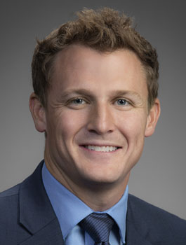 A man with short, light brown hair is smiling. He is wearing a blue shirt, a blue suit jacket, and a dark tie. The background is a plain gray gradient.