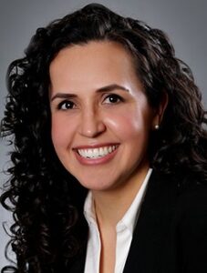 Smiling person with long, curly dark hair, wearing a black blazer and white shirt, against a gray background.