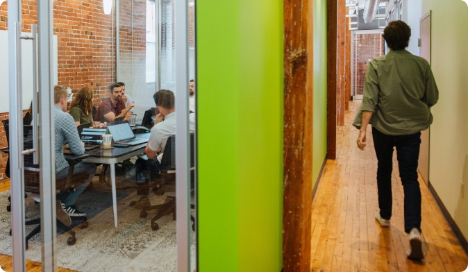 A person walks down a hallway with wooden floors and green walls. On the left, a group of people are seated at a rectangular table in a glass-walled meeting room, engaged in discussion, with laptops open.
