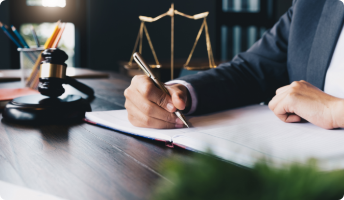 A person in a suit writes in a notebook at a wooden desk. A judges gavel and scales of justice are visible, suggesting a legal or professional setting.