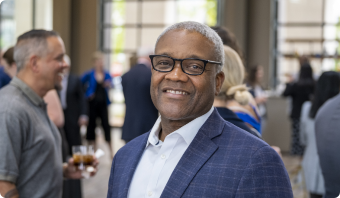 A man in a blue suit and glasses smiles at the camera in a social setting. Other people are mingling in the blurred background, holding drinks. The event appears to be indoors with large windows.