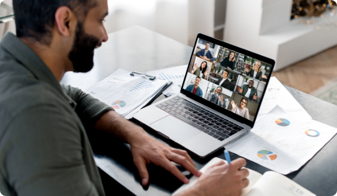 A man is sitting at a desk with charts and documents, participating in a video call on a laptop. The screen shows multiple people in a grid layout. He is holding a pen, ready to take notes.