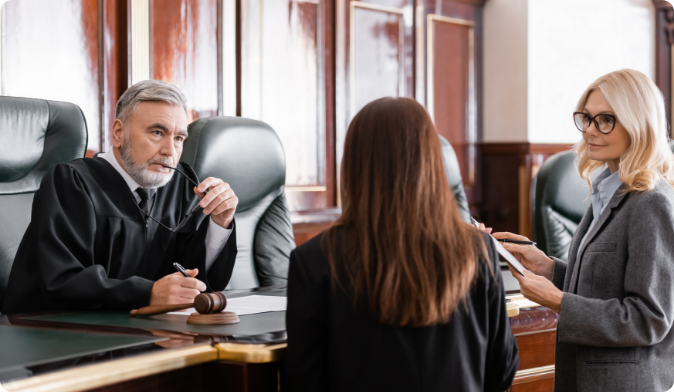 A judge in black robes sits at the bench with a thoughtful expression, holding glasses. A lawyer stands beside him, holding documents, and a woman with long hair stands facing the judge. The courtroom has wooden paneling.